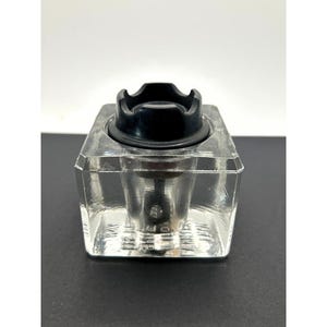 May include: A clear glass inkwell with a square base and a black, ridged top. The inkwell has a classic design, suitable for calligraphy or vintage desk decor. The inkwell is empty, showing its transparent nature.