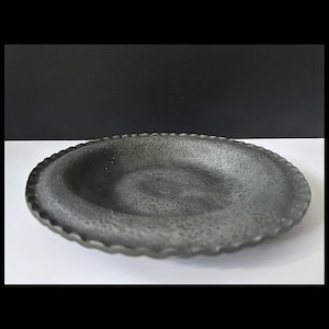 May include: A dark grey, round, shallow dish with a textured surface and a scalloped edge. The dish is centred on a white surface with a black background.