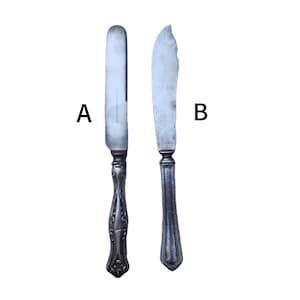 May include: Two antique butter knives with silver-colored blades and ornate handles. Knife A has a rounded blade and a detailed handle. Knife B has a curved blade and a simpler handle design. Both knives are set against a white background.