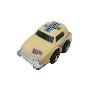 May include: A small, cream-coloured toy car with black wheels and a silver front bumper. The car features the word "Racing" on the front and "354GT" on the roof, with blue and pink accents. A vintage toy car.
