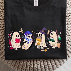 May include: Black t-shirt featuring a whimsical design of four cartoon ghosts, each holding items like books, a wand, and a broom. The ghosts are in various poses, with colorful accents. The shirt is made of a soft material.