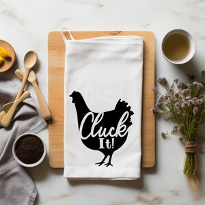 May include: White kitchen towel with a black silhouette of a chicken and the words "Cluck It!" in a whimsical font. The towel is on a wooden cutting board, surrounded by wooden spoons, a bowl of seeds, and a cup of liquid.