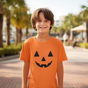 May include: An orange t-shirt with a black jack-o'-lantern face design. The shirt is short-sleeved and worn by a person smiling in an outdoor setting. The background is blurred with palm trees and a walkway.