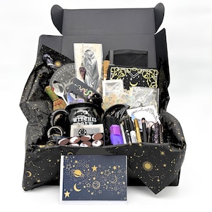 May include: A black gift box filled with various items including a black mug with the text Witches Brew, a purple crystal pendulum, a small broom, pens, markers, a card with celestial patterns, and black tissue paper with gold star and moon prints.