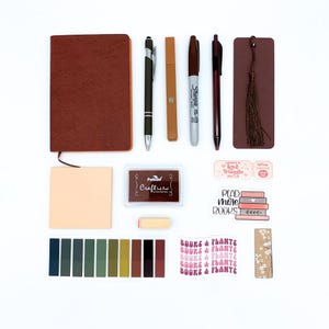 May include: An assortment of stationery items, including a brown leather-bound notebook, pens, a Sharpie marker, a bookmark with a tassel, and stickers with the text "Read More Books" and "Books & Plants".