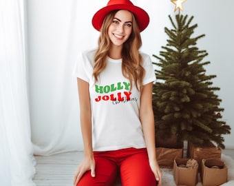 May include: A woman wearing a white t-shirt with the words "HOLLY JOLLY Christmas" in green, red, and black. She is also wearing red pants and a red hat. A small Christmas tree is in the background.