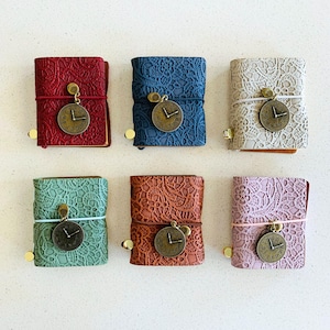May include: Six small, decorative journals in various colors: red, blue, white, green, brown, and pink. Each journal features a lace cover, a clock charm, and a matching elastic band closure. The journals are arranged on a white surface.
