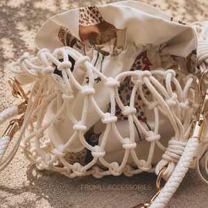 May include: A drawstring bucket bag featuring a cream-colored fabric pouch with a multi-colored chicken print, encased in a white knotted macrame net. The bag includes a braided white shoulder strap with gold-tone hardware and the text FROMLI_ACCESSORIES.