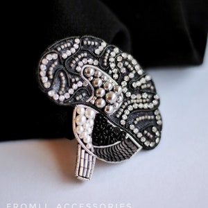 Könnte beinhalten: Handgefertigte Gehirn-Brosche mit aufwendiger Perlenarbeit. Das Design umfasst schwarze, silberne und weiße Perlen, Pailletten und Perlen. Der Text "FROMLI_ACCESSORIES" ist sichtbar. Die Brosche ist vor schwarzem Stoff platziert.