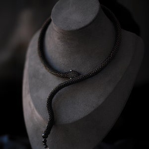 Puede incluir: Un collar de cuentas negras con forma de serpiente, exhibido en un maniquí de busto gris.