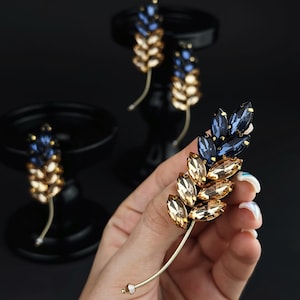 Puede incluir: Primer plano de un conjunto de broche y pendientes dorados. El broche y los pendientes están diseñados con un estilo de espiga de trigo, con detalles de cristal azul y ámbar. El conjunto se muestra sobre un fondo negro.