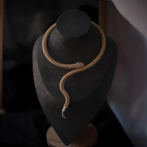 Könnte beinhalten: Eine goldfarbene Schlangenkette, präsentiert auf einer schwarzen Schmuckbüste. Die Halskette besteht aus kleinen, strukturierten Perlen, mit einem Schlangenkopf-Design und einem Anhänger mit kleinen weißen Perlen. Die Büste steht vor einem dunklen Hintergrund.