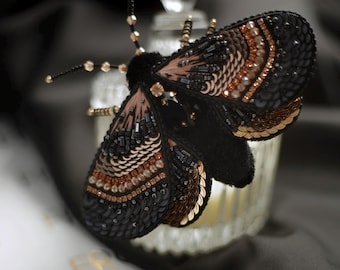 May include: A detailed, black moth brooch with intricate bead and sequin detailing in shades of gold and brown. The moth's wings are adorned with layers of sequins and beads, creating a textured and elegant accessory.