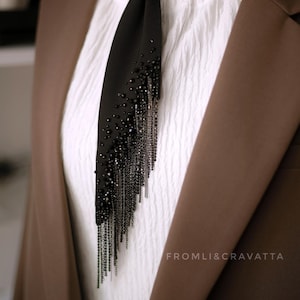 Puede incluir: Una corbata negra con una larga cadena de cuentas negras y detalles plateados. La corbata se lleva con una camisa blanca y una chaqueta marr&oacute;n. El texto "FROMLI&CRAVATTA" es visible en la esquina inferior derecha de la imagen.