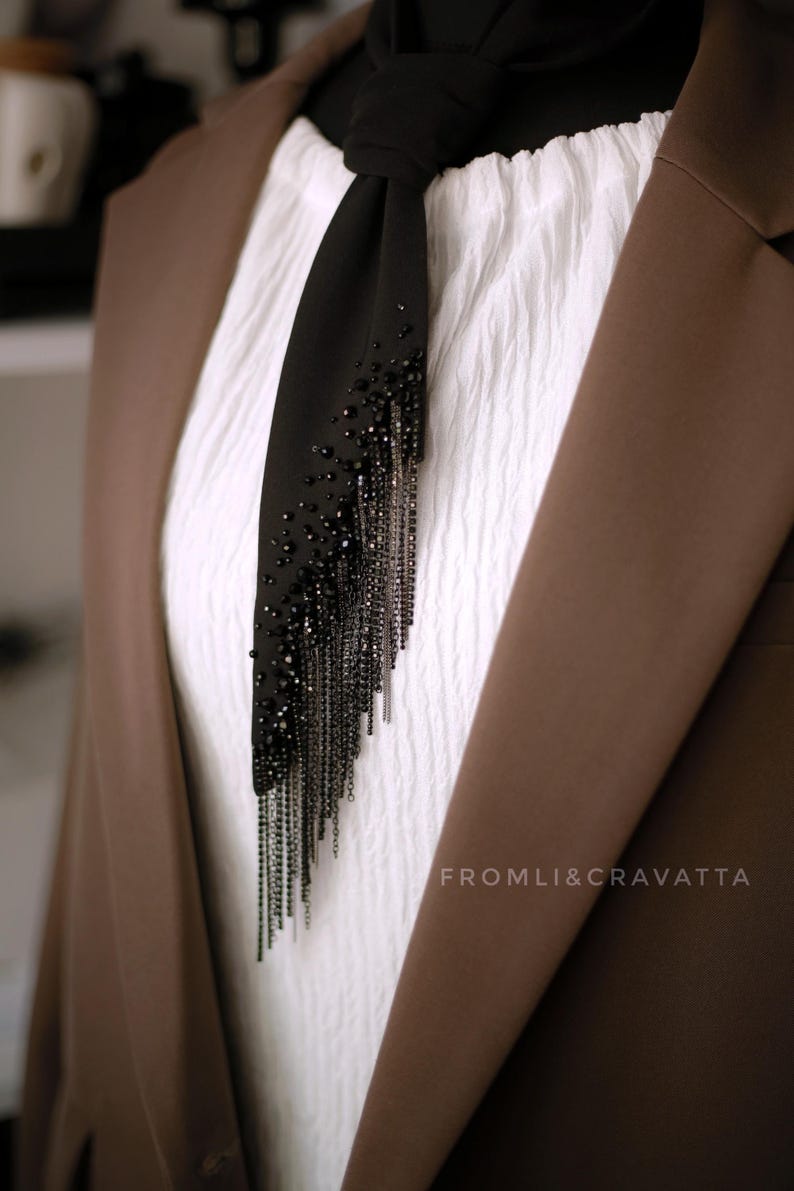 Puede incluir: Una corbata negra con una larga cadena de cuentas negras y detalles plateados. La corbata se lleva con una camisa blanca y una chaqueta marr&oacute;n. El texto "FROMLI&CRAVATTA" es visible en la esquina inferior derecha de la imagen.