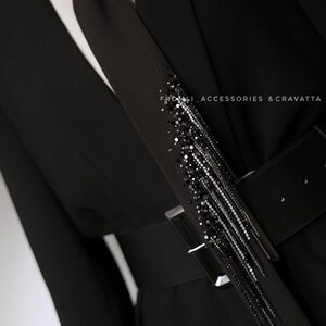 Puede incluir: Un blazer negro con una corbata negra que presenta un largo detalle de flecos con cuentas. El blazer tiene un cintur&oacute;n negro con una hebilla plateada. El texto "FROM.LI ACCESSORIES & CRAVATTA" es visible en la imagen.