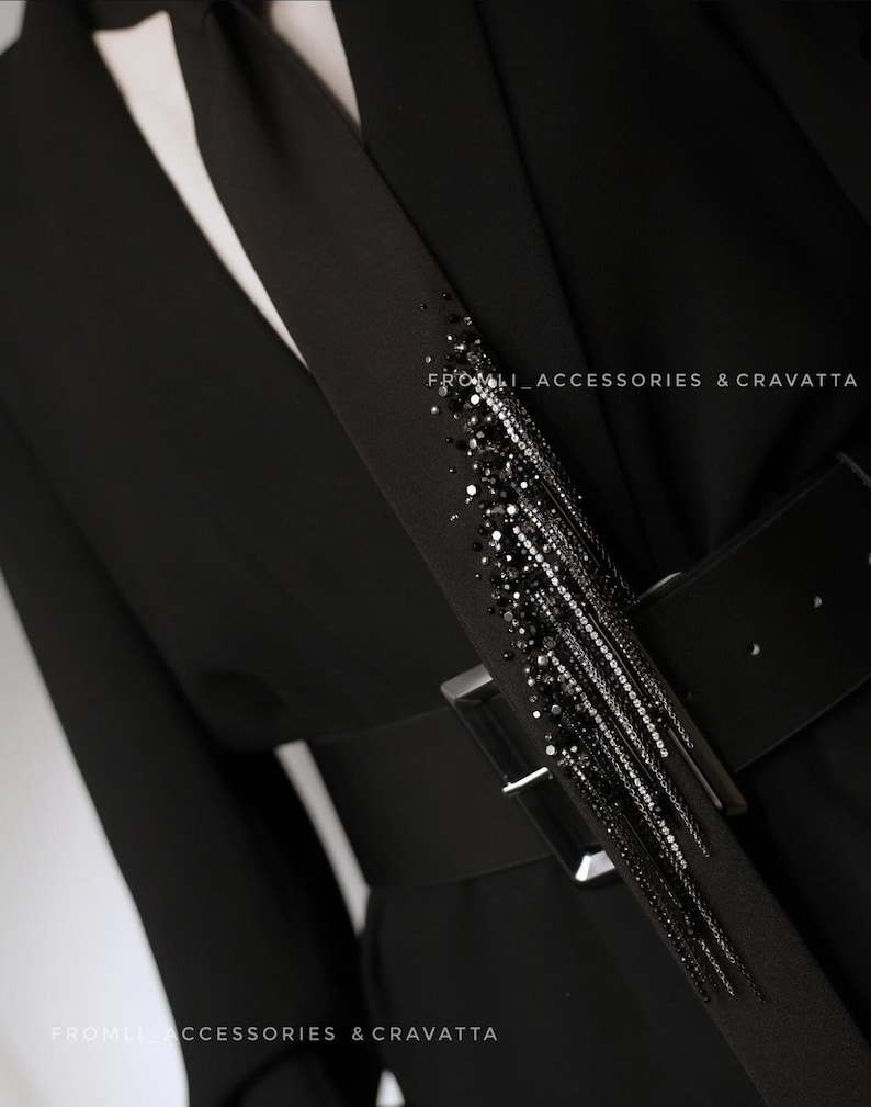 Puede incluir: Un blazer negro con una corbata negra que presenta un largo detalle de flecos con cuentas. El blazer tiene un cintur&oacute;n negro con una hebilla plateada. El texto "FROM.LI ACCESSORIES & CRAVATTA" es visible en la imagen.