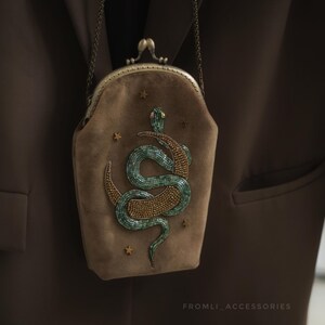 Könnte beinhalten: Eine braune Wildleder-Clutch mit Metallverschluss und Kette. Die Clutch hat ein gesticktes Design einer grünen und goldenen Schlange und mehrere goldene Sterne. Der Text "FROMLI_ACCESSORIES" ist unten rechts sichtbar.