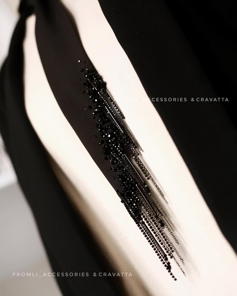 Puede incluir: Una corbata negra con un detalle de flecos de cuentas. El fleco est&aacute; hecho de peque&ntilde;as cuentas negras y cadenas met&aacute;licas. El texto "FROMLI_ACCESSORIES & CRAVATTA" es visible en la parte inferior de la imagen.