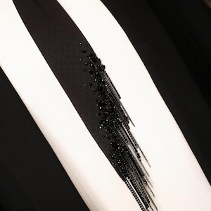 Puede incluir: Una tela a rayas blancas y negras con un detalle de flecos negros con cuentas. La tela es probablemente una prenda de vestir, como una camisa o una chaqueta.