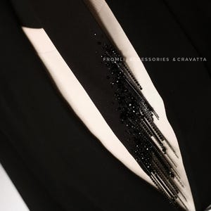 Puede incluir: Un blazer negro con una camisa blanca debajo. Un adorno negro de cuentas y cadena est&aacute; unido a la solapa del blazer. El texto "FROMLI ACCESSORIES & CRAVATTA" es visible en el blazer.
