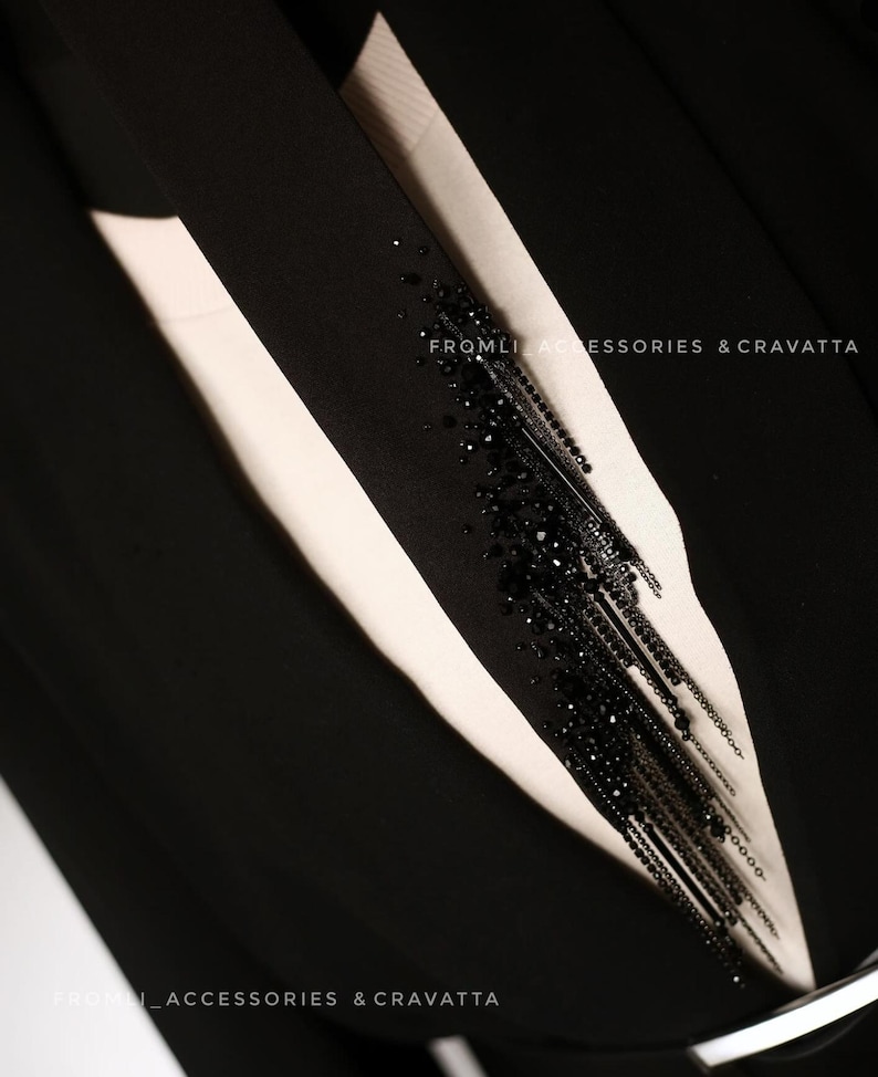 Puede incluir: Un blazer negro con una camisa blanca debajo. Un adorno negro de cuentas y cadena est&aacute; unido a la solapa del blazer. El texto "FROMLI ACCESSORIES & CRAVATTA" es visible en el blazer.