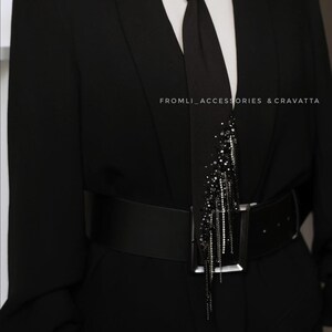 Puede incluir: Un blazer negro con una corbata negra y un cintur&oacute;n negro. La corbata tiene un adorno de cuentas negras y plateadas. El texto "FROMLI_ACCESSORIES & CRAVATTA" es visible en el blazer.
