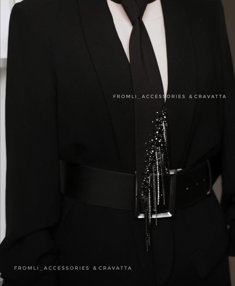 Puede incluir: Un blazer negro con una corbata negra y un cintur&oacute;n negro. La corbata tiene un adorno de cuentas negras y plateadas. El texto "FROMLI_ACCESSORIES & CRAVATTA" es visible en el blazer.
