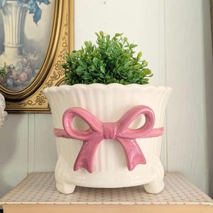 May include: A cream-colored ceramic planter with a scalloped rim and a raised, glossy pink bow detail wrapped around the center. The planter features three small, rounded feet at the base and contains a small, vibrant green artificial plant.