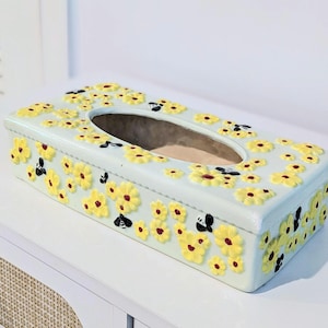 May include: A rectangular light blue tissue box cover featuring raised yellow flowers with dark centers and small black and white bees scattered across the top and sides. The box has an oval opening in the center for dispensing tissues.