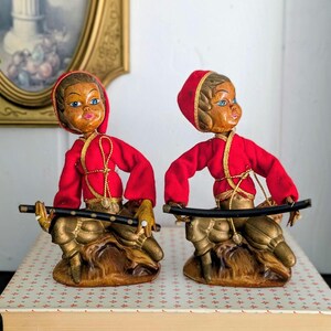 May include: Two vintage figurines depicting figures with brown skin, blue eyes, and pink lips, wearing red hooded jackets with gold trim and gold pants. Each figure sits on a brown base and holds a long, thin black rod across their lap.