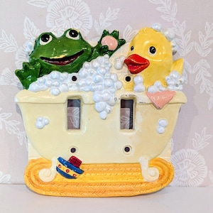 May include: A double light switch plate featuring a sculpted bathtub scene with a green frog and a yellow duck surrounded by white bubbles. The bathtub is cream-colored with a textured orange base and a small blue and red toy boat attached to the front.