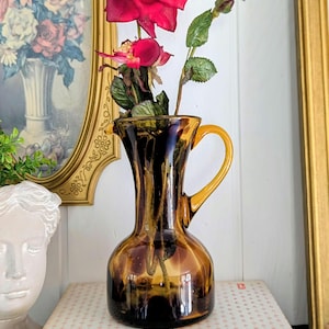 May include: A translucent amber-colored glass pitcher with a rounded base, a slender neck, and a curved handle. The pitcher features a dark brown tortoiseshell-like pattern throughout the glass and contains a stem with two deep red artificial roses.