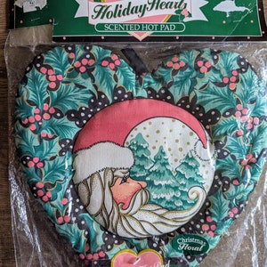 May include: Victorian Holiday Hearts scented hot pad in the shape of a heart with a Santa Claus design and a green holly pattern. The hot pad is packaged in a clear plastic bag with a pink and green label that says "Scented Hot Pad".
