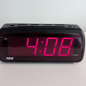 May include: A black digital alarm clock displaying the time 4:08 PM in bright pink numerals. The clock has a rectangular shape with a curved front and the brand name "TDE" is visible.