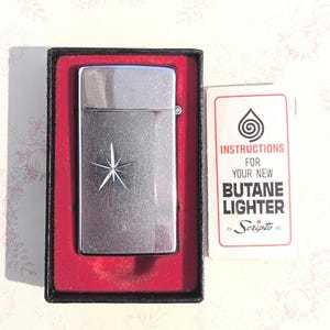 May include: A silver-colored butane lighter with a starburst design, presented in a black box with a red interior. Includes a card with instructions for the lighter. The card reads "Instructions for your new Butane Lighter by Scripto Inc."