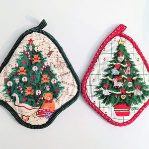 May include: Two diamond-shaped quilted pot holders featuring Christmas tree illustrations. The left one has a green border, a goose, and teddy bears. The right one has a red border, a grid pattern, and various ornaments on the tree.