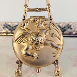 May include: A gold-colored compact mirror with an embossed cherub design, set on a decorative stand. The compact features floral accents and is embellished with small, sparkling rhinestones. The stand has a bamboo-like design.