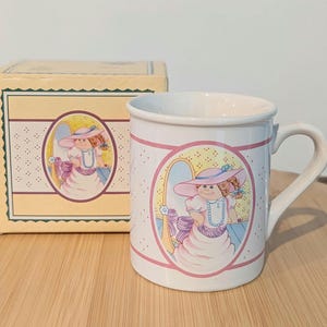 May include: White ceramic mug with a pink and purple illustration of a doll wearing a hat and dress. The mug has a matching box with the same illustration. The mug has a handle and pink trim.