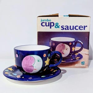 May include: A dark blue jumbo cup and saucer set featuring a painted moon face with a purple crescent, yellow stars, and colorful dots. The packaging box displays the text jumbo cup & saucer and natural wonders, matching the cup's design.
