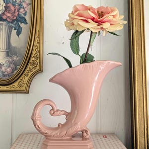 May include: A pink ceramic cornucopia vase with a curved handle and decorative base. A large, light pink and yellow artificial rose is placed inside the vase. The vase is displayed on a patterned surface, with a framed painting in the background.
