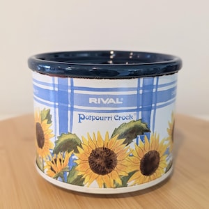 May include: A cylindrical Rival Potpourri Crock with a dark blue rim. The exterior features a white and blue plaid pattern decorated with yellow sunflowers and green leaves. The text RIVAL and Potpourri Crock are printed on the front surface.