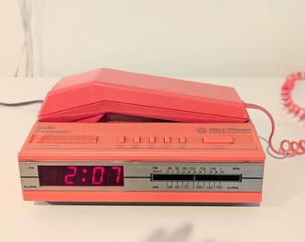 May include: A coral pink Radio Roommate alarm clock radio with a matching telephone handset resting on top. The device features a digital display showing 2:07, an analog AM/FM radio tuner, and a coiled cord. The front panel includes the text Radio Roommate and Bell Phones.