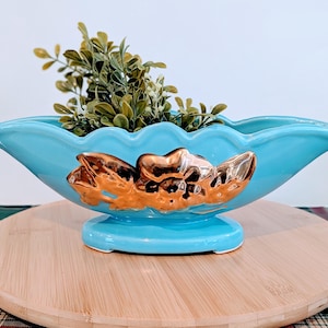 May include: A turquoise ceramic planter with a gold floral design. The planter is filled with green faux foliage and sits on a round wooden surface. The planter has a scalloped rim and a pedestal base.