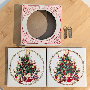May include: Two square ceramic trivets with a Christmas tree design, a box labeled "Ceramic Tile Trivets", and two metal hangers. The trivets feature a Christmas tree with red ribbon, candles, and toys. The box is white with red text.