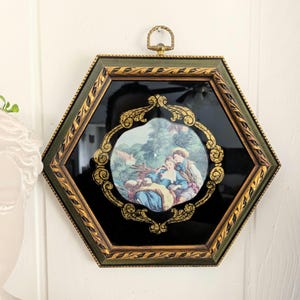 May include: A hexagonal wall hanging with a romantic scene. The frame is green and gold, surrounding a black background. The central image depicts a couple in a garden setting, framed by ornate gold detailing. A brass loop is at the top for hanging.