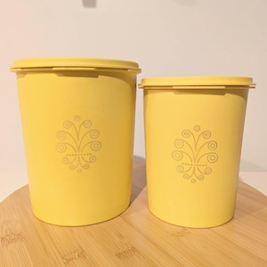 May include: Two yellow plastic canisters with lids. Each canister has a decorative floral design. The canisters are different sizes and sit on a wooden surface. The canisters are likely for kitchen storage.