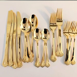 May include: A set of gold-colored stainless steel flatware including four knives, four spoons, and six forks. Each piece features a polished reflective finish and a decorative fan-shaped motif at the base of the handle, arranged in a group.