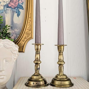 May include: Two antique brass candlesticks with gray taper candles. The candlesticks have a detailed, ornate design and are placed on a patterned surface. A framed painting and a white bust are in the background.