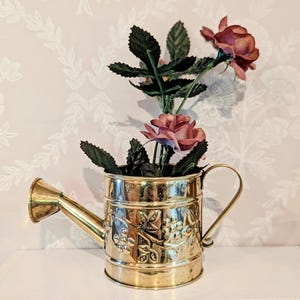 May include: A small, decorative brass watering can with embossed floral designs, holding faux pink roses and green leaves. The watering can has a spout and a curved handle. The background is a light pink wall with a white floral pattern.